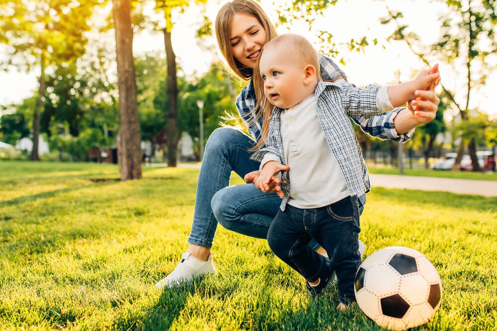 Happy young mom and her little son play soccer together outdoors in the park